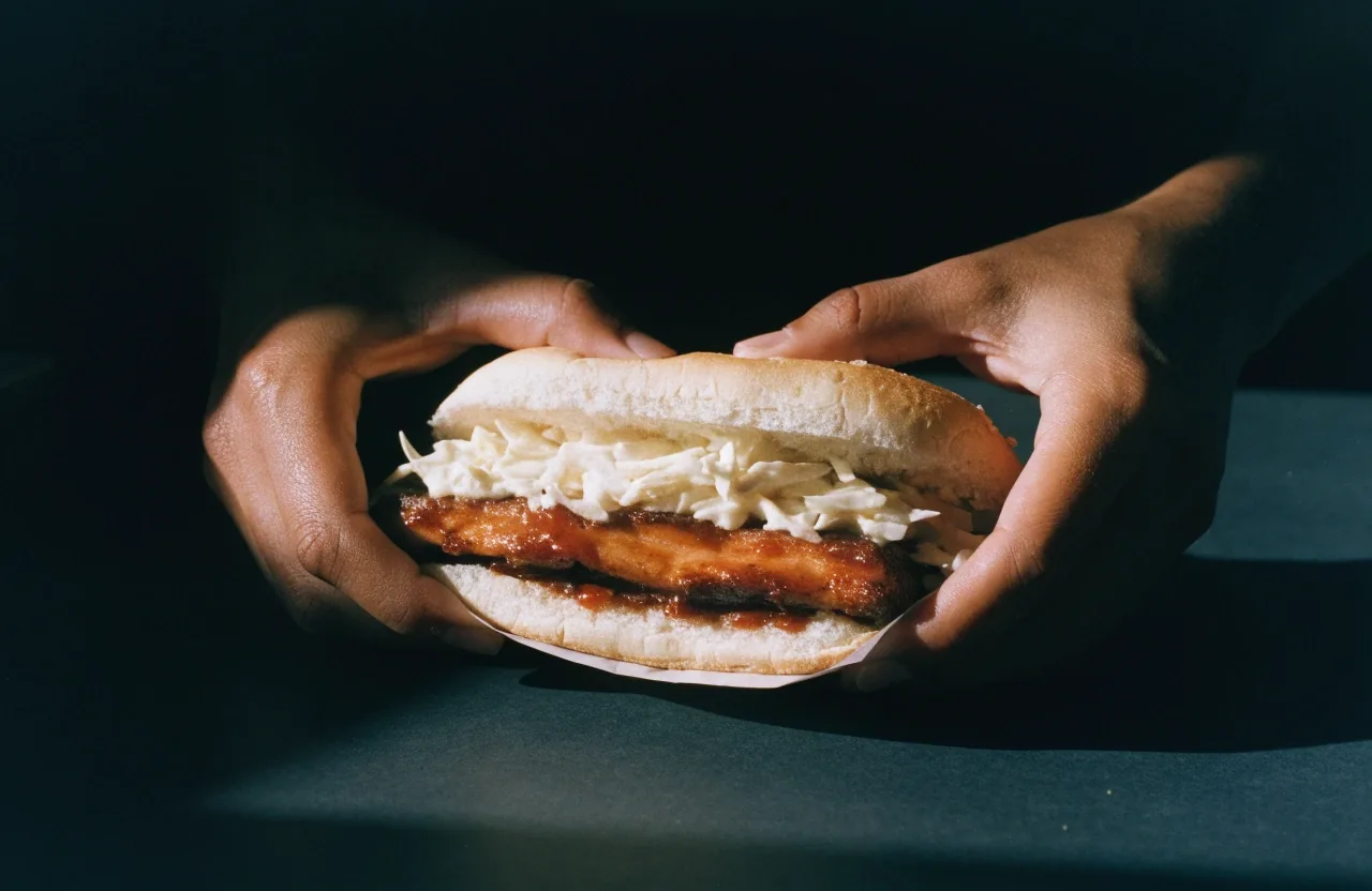 Hands pulling a slaw-topped rib sandwich from plain wrapper paper, meat and slaw texture visible at the bun edge